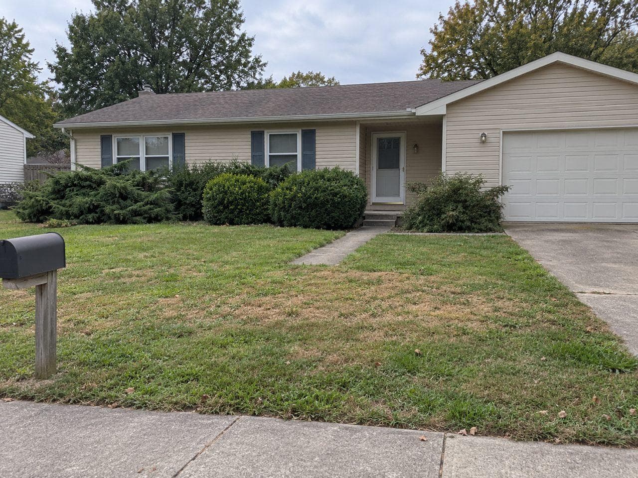 A single-story beige ranch house with blue shutters and a white garage door, overgrown junipers and boxwoods under the windows, a patchy lawn and a short concrete walk